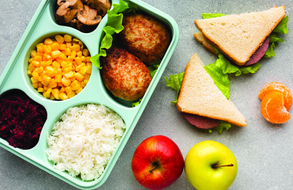 An overhead photo of a school lunch that includes a tray of cooked items, a sandwich and fruit